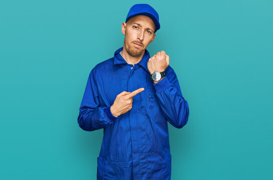 Bald man with beard wearing builder jumpsuit uniform in hurry pointing to watch time, impatience, looking at the camera with relaxed expression