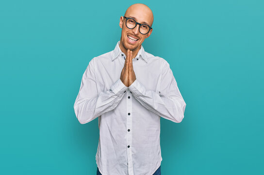 Bald man with beard wearing business shirt and glasses praying with hands together asking for forgiveness smiling confident.