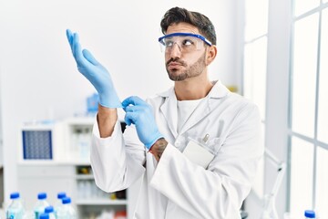 Young hispanic man working at scientist laboratory putting gloves on smiling looking to the side and staring away thinking.