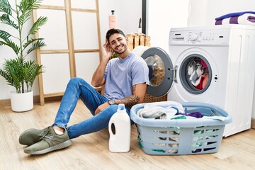 Young hispanic man putting dirty laundry into washing machine smiling with hand over ear listening an hearing to rumor or gossip. deafness concept.