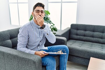 Young psychologist man at consultation office laughing and embarrassed giggle covering mouth with hands, gossip and scandal concept