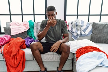 Young african american man sitting on the sofa with dirty laundry clothes smelling something stinky...