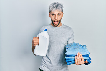 Young hispanic man with modern dyed hair holding jeans for laundry and detergent bottle afraid and shocked with surprise and amazed expression, fear and excited face.