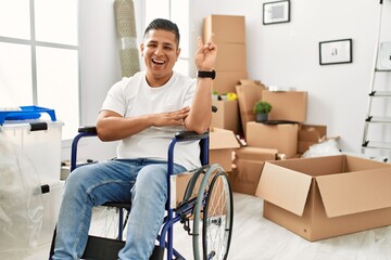 Young hispanic man sitting on wheelchair at new house smiling with happy face winking at the camera...