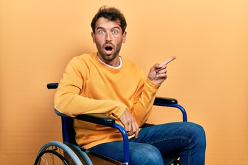 Handsome man with beard sitting on wheelchair surprised pointing with finger to the side, open...