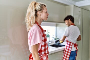 Young caucasian woman wearing apron and husband doing housework washing dishes looking to side,...