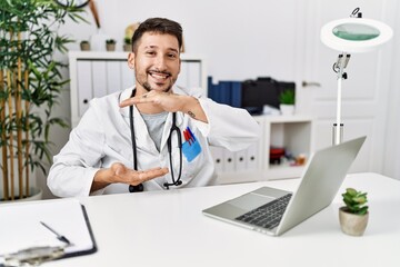 Young doctor working at the clinic using computer laptop gesturing with hands showing big and large size sign, measure symbol. smiling looking at the camera. measuring concept.
