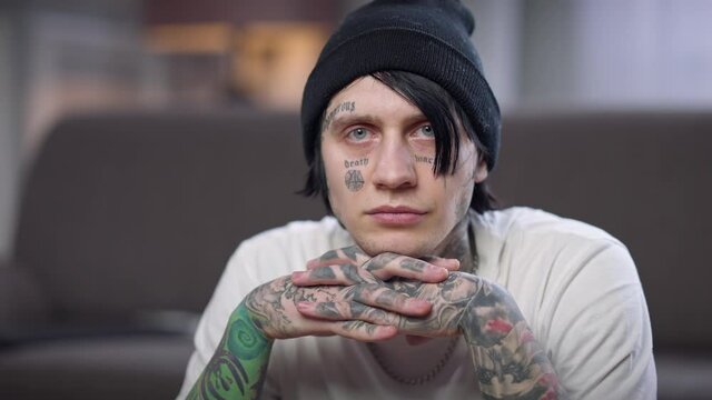 Portrait of man with tattooed face and hands turning looking at camera with thoughtful facial expression. Young Caucasian guy posing indoors at home in living room
