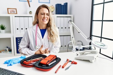 Young beautiful doctor woman with reflex hammer and medical instruments with hand on stomach...