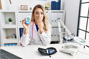 Young beautiful doctor woman holding glucose meter covering mouth with hand, shocked and afraid for...