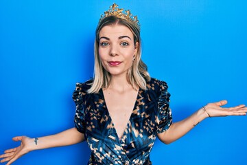 Beautiful caucasian woman wearing princess crown clueless and confused expression with arms and hands raised. doubt concept.