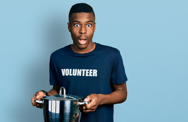 Young african american man wearing volunteer holding cooking pot afraid and shocked with surprise...