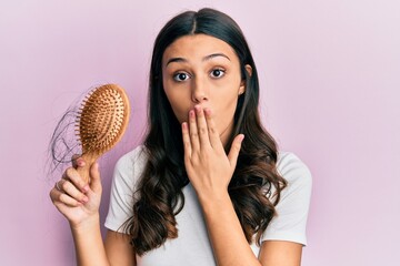 Young hispanic woman styling hair using comb covering mouth with hand, shocked and afraid for...