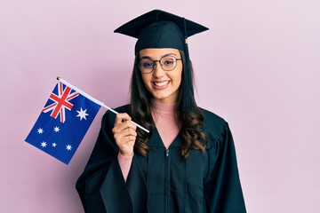 Young hispanic woman wearing graduation uniform holding australia flag looking positive and happy standing and smiling with a confident smile showing teeth