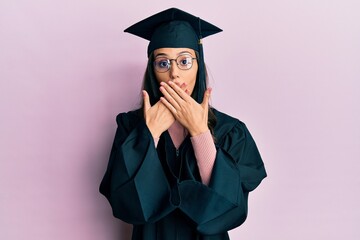 Young hispanic woman wearing graduation cap and ceremony robe shocked covering mouth with hands for...