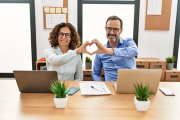 Middle age hispanic woman and man sitting with laptop at the office smiling in love doing heart...