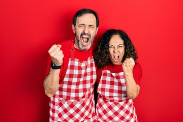 Middle age couple of hispanic woman and man wearing cook apron angry and mad raising fist frustrated and furious while shouting with anger. rage and aggressive concept.