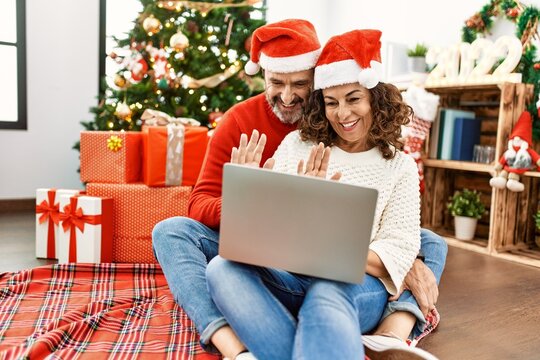 Middle age hispanic couple wearing christmas hat. Sitting on the floor having video call using laptop at home.