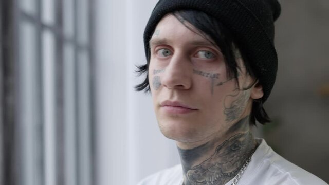 Side view young man looking out window turning tattooed face looking at camera. Close-up portrait of Caucasian guy posing indoors at home