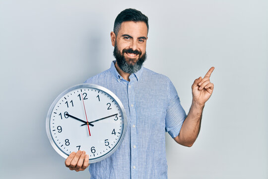 Young hispanic man holding big clock smiling happy pointing with hand and finger to the side