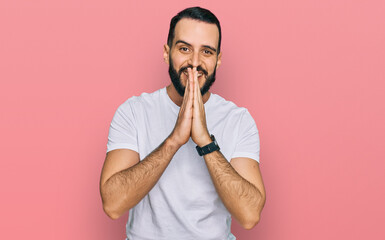 Young man with beard wearing casual white t shirt praying with hands together asking for forgiveness smiling confident.