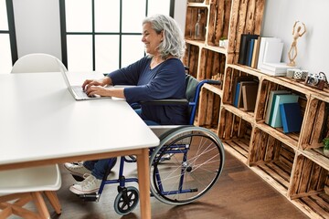 Middle age grey-haired disabled woman using laptop sitting on wheelchair at home.