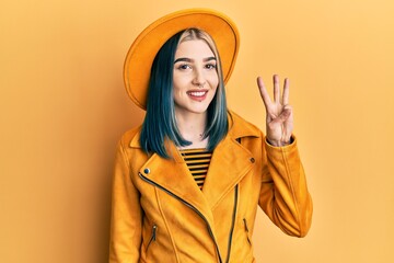 Young modern girl wearing yellow hat and leather jacket showing and pointing up with fingers number three while smiling confident and happy.