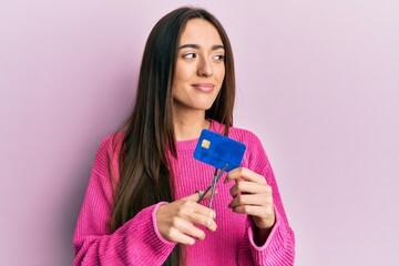 Young hispanic girl cutting credit card using scissors smiling looking to the side and staring away...