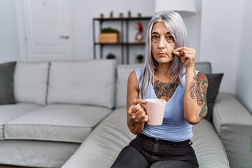 Middle age grey-haired woman drinking coffee sitting on the sofa at home mouth and lips shut as zip...
