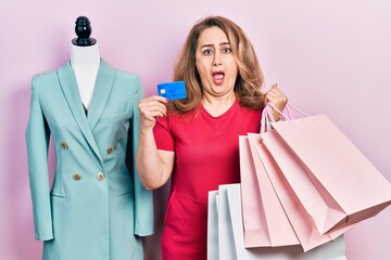 Middle age caucasian woman holding shopping bags and credit card in shock face, looking skeptical and sarcastic, surprised with open mouth