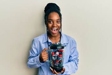African american woman with braided hair holding food processor mixer machine with fruits celebrating crazy and amazed for success with open eyes screaming excited.