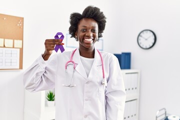 African doctor woman holding purple ribbon awareness at medical clinic looking positive and happy standing and smiling with a confident smile showing teeth