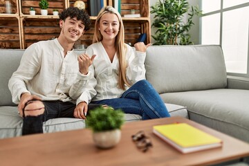 Young beautiful couple sitting on the sofa at home pointing thumb up to the side smiling happy with open mouth