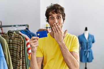 Young hispanic man at retail shop holding credit card covering mouth with hand, shocked and afraid...