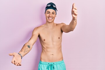 Young hispanic man wearing swimwear and swimmer glasses looking at the camera smiling with open arms for hug. cheerful expression embracing happiness.