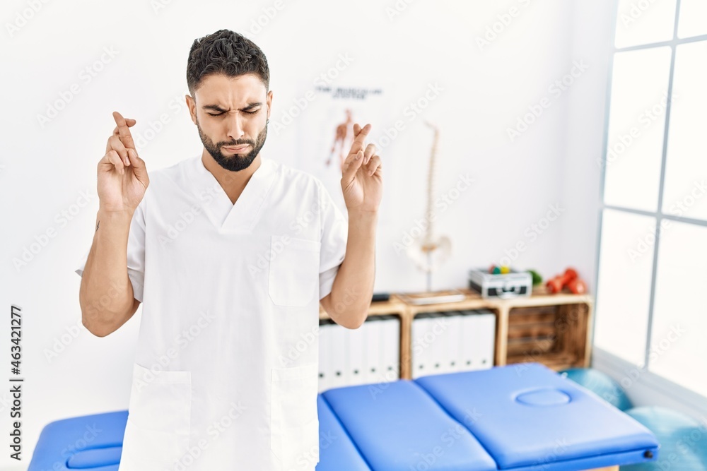 Wall mural Young handsome man with beard working at pain recovery clinic gesturing finger crossed smiling with hope and eyes closed. luck and superstitious concept.