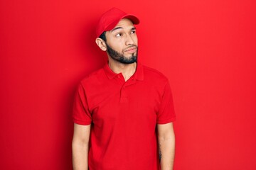 Hispanic man with beard wearing delivery uniform and cap smiling looking to the side and staring away thinking.