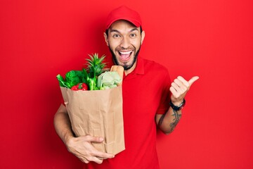 Hispanic man with beard wearing courier uniform with groceries from supermarket pointing thumb up...