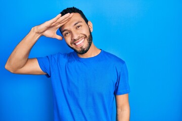 Naklejka premium Hispanic man with beard wearing casual blue t shirt very happy and smiling looking far away with hand over head. searching concept.