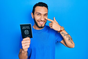 Hispanic man with beard holding italy passport pointing with hand finger to face and nose, smiling...