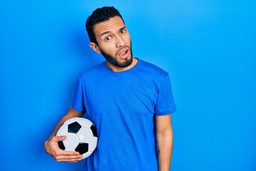 Hispanic man with beard holding soccer ball in shock face, looking skeptical and sarcastic, surprised with open mouth