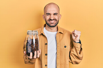 Young bald man holding jar of raisins screaming proud, celebrating victory and success very excited with raised arm