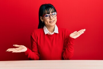 Young brunette woman with bangs wearing glasses sitting on the table clueless and confused...