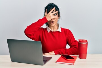 Young brunette woman with bangs working at the office with laptop peeking in shock covering face...