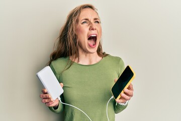 Young blonde woman charging smartphones angry and mad screaming frustrated and furious, shouting with anger looking up.
