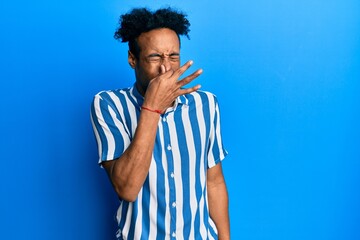 Young african american man with beard wearing casual striped shirt smelling something stinky and...