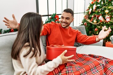 Young hispanic woman surprisng boyfriend with valentine gift sitting on the sofa by christmas tree at home.