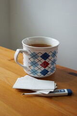 Mug of tea with a checkered pattern, paper handkerchiefs and a personal thermometer on a wooden table