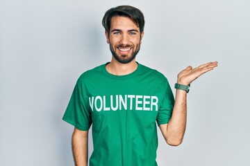 Young hispanic man wearing volunteer t shirt smiling cheerful presenting and pointing with palm of...