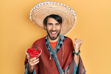 Young hispanic man wearing mexican hat holding chili screaming proud, celebrating victory and...